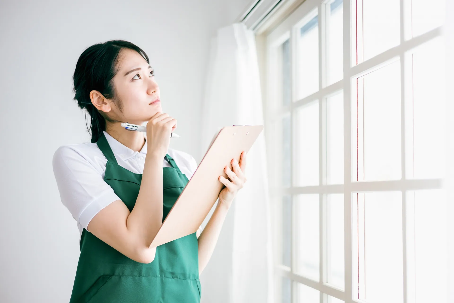 アルミサッシをDIYで塗装するのは可能か、窓の前で思案している緑のエプロンをした女性