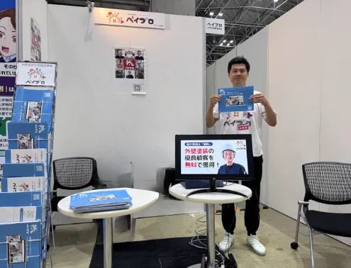業界展示会のブースで担当者がチラシを持って立っている写真。